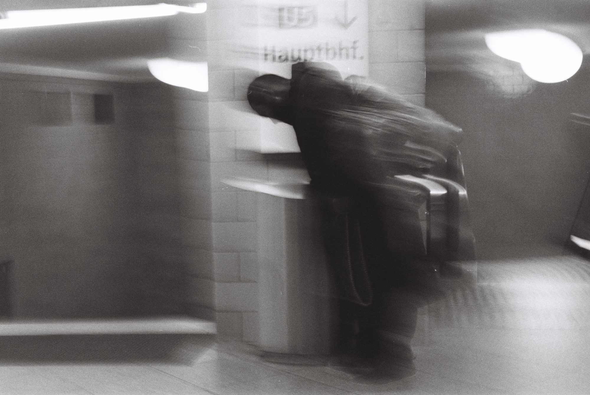 Blurred black and white figure leaning against a tiled pillar under a Hauptbahnhof sign