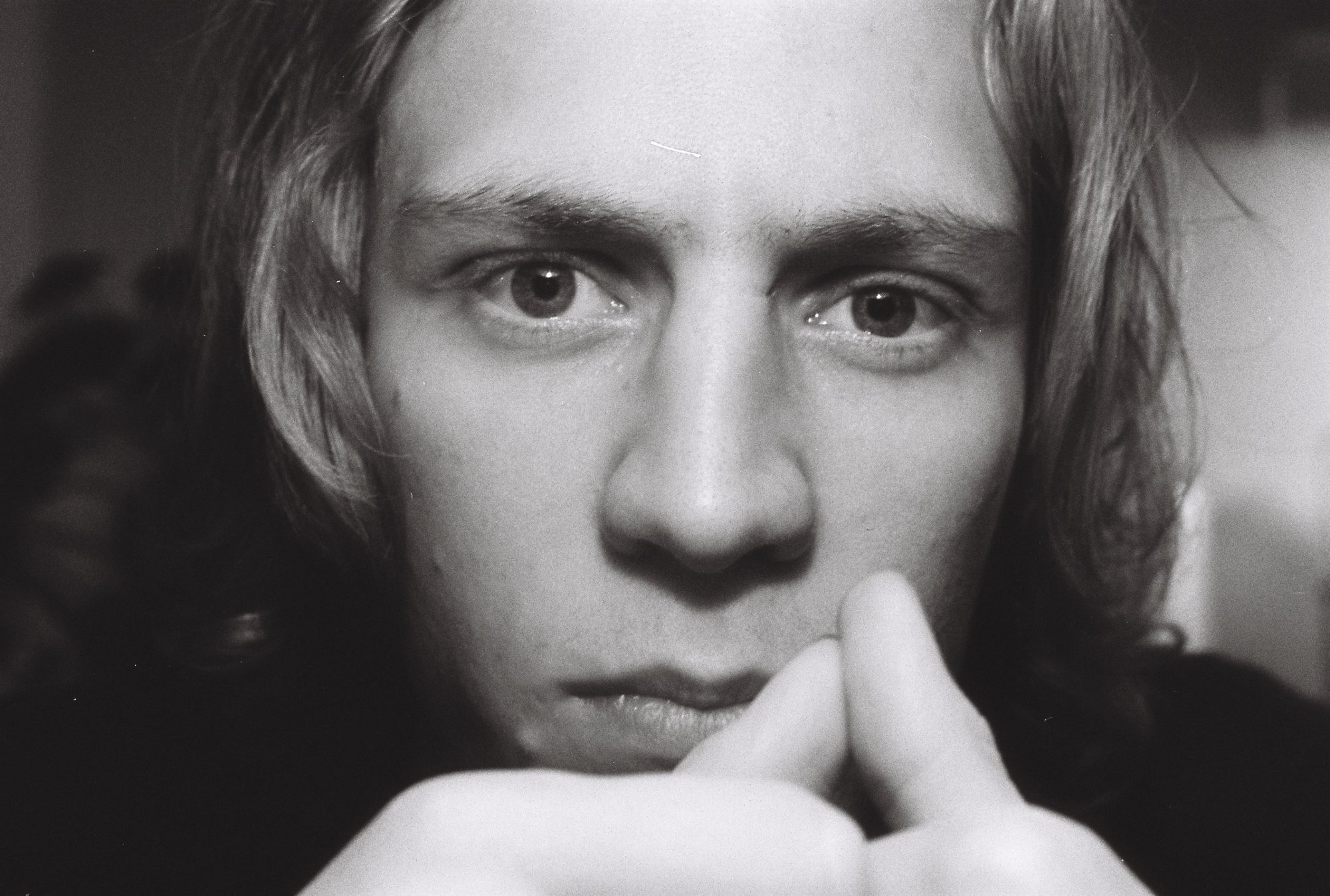 Close black and white portrait of a young man with long hair, hands at his chin