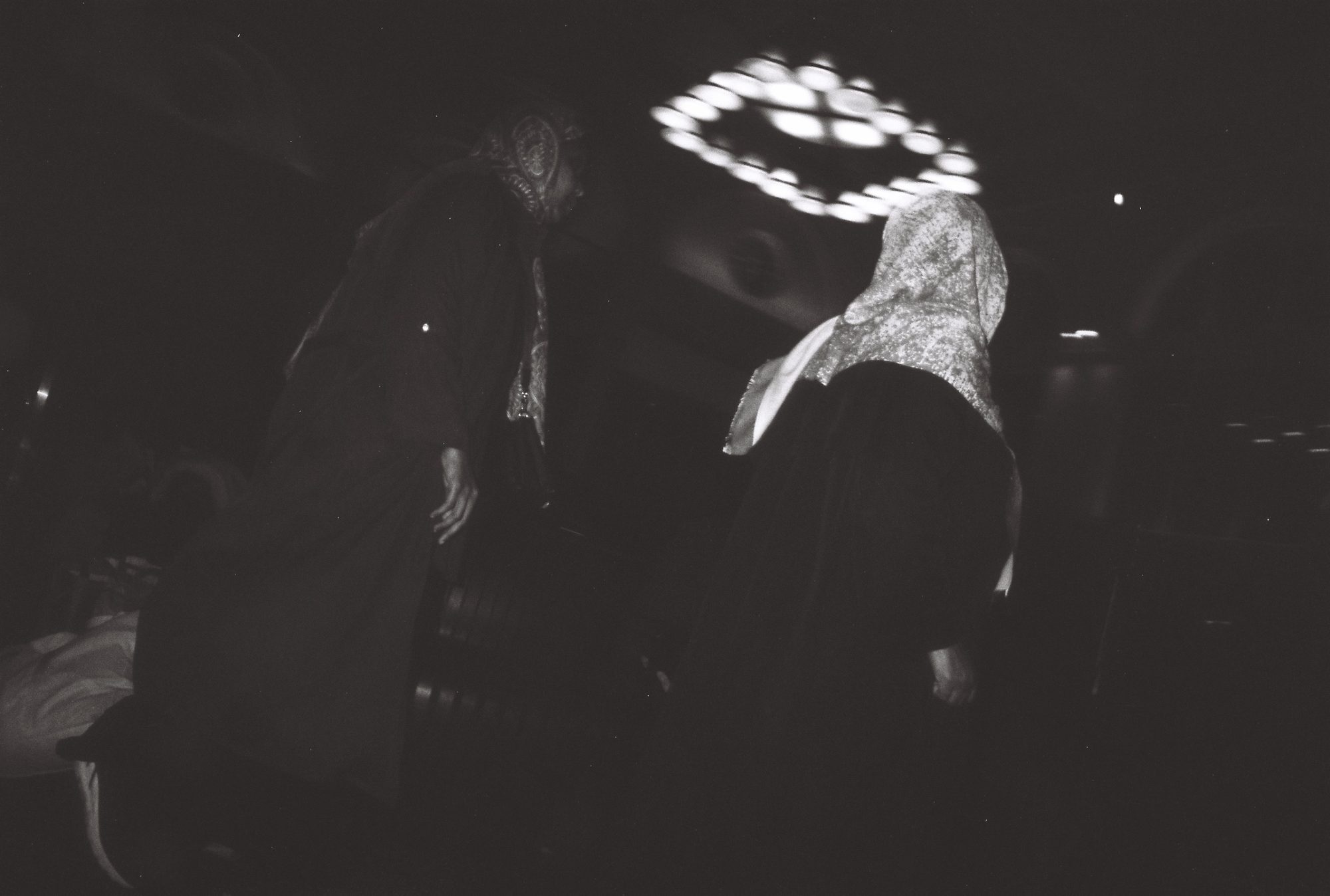 Dark night scene of two figures in patterned headscarves under a blurred ring of lights