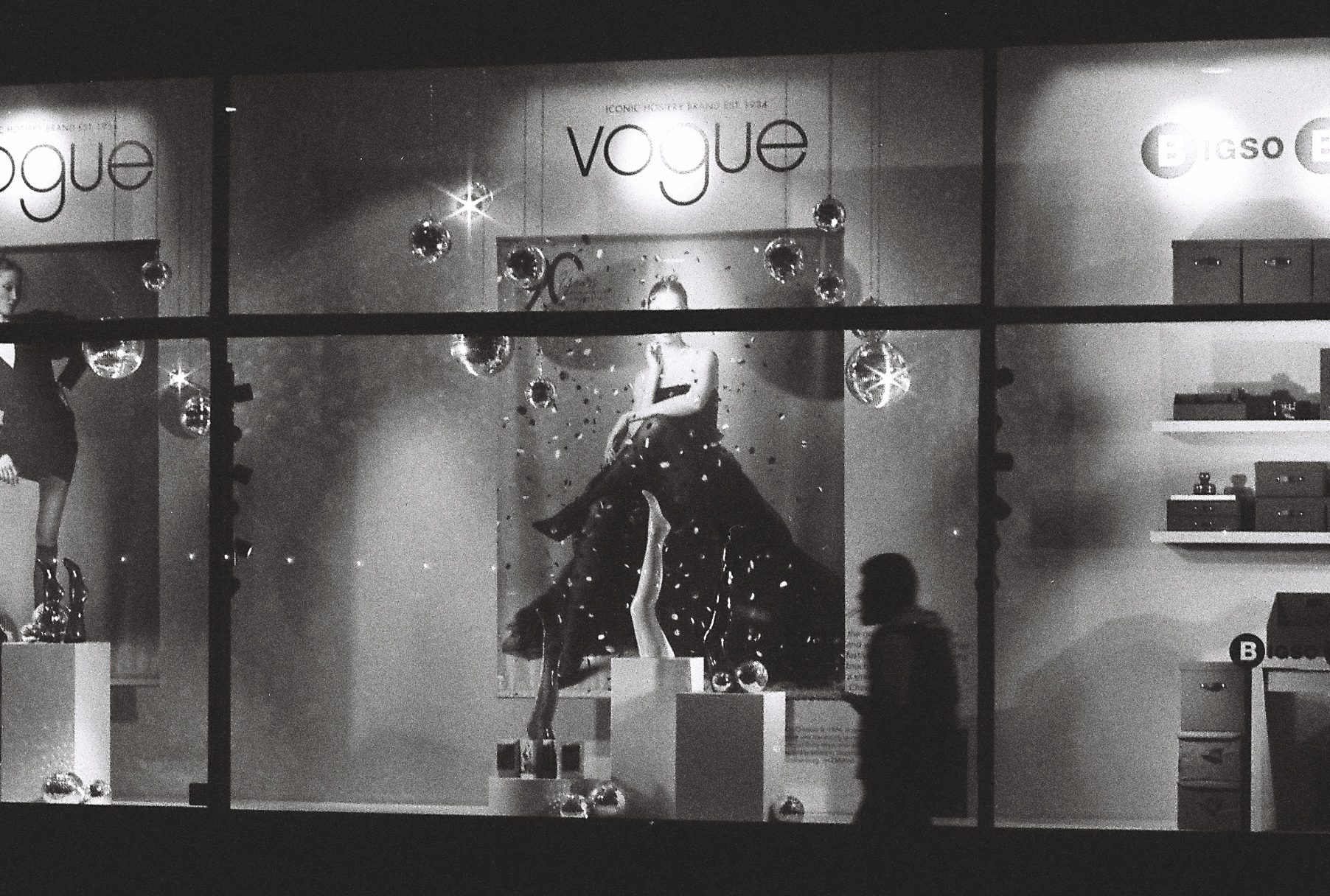 Black and white night photo of a lit shop window, a silhouetted passer-by walking past
