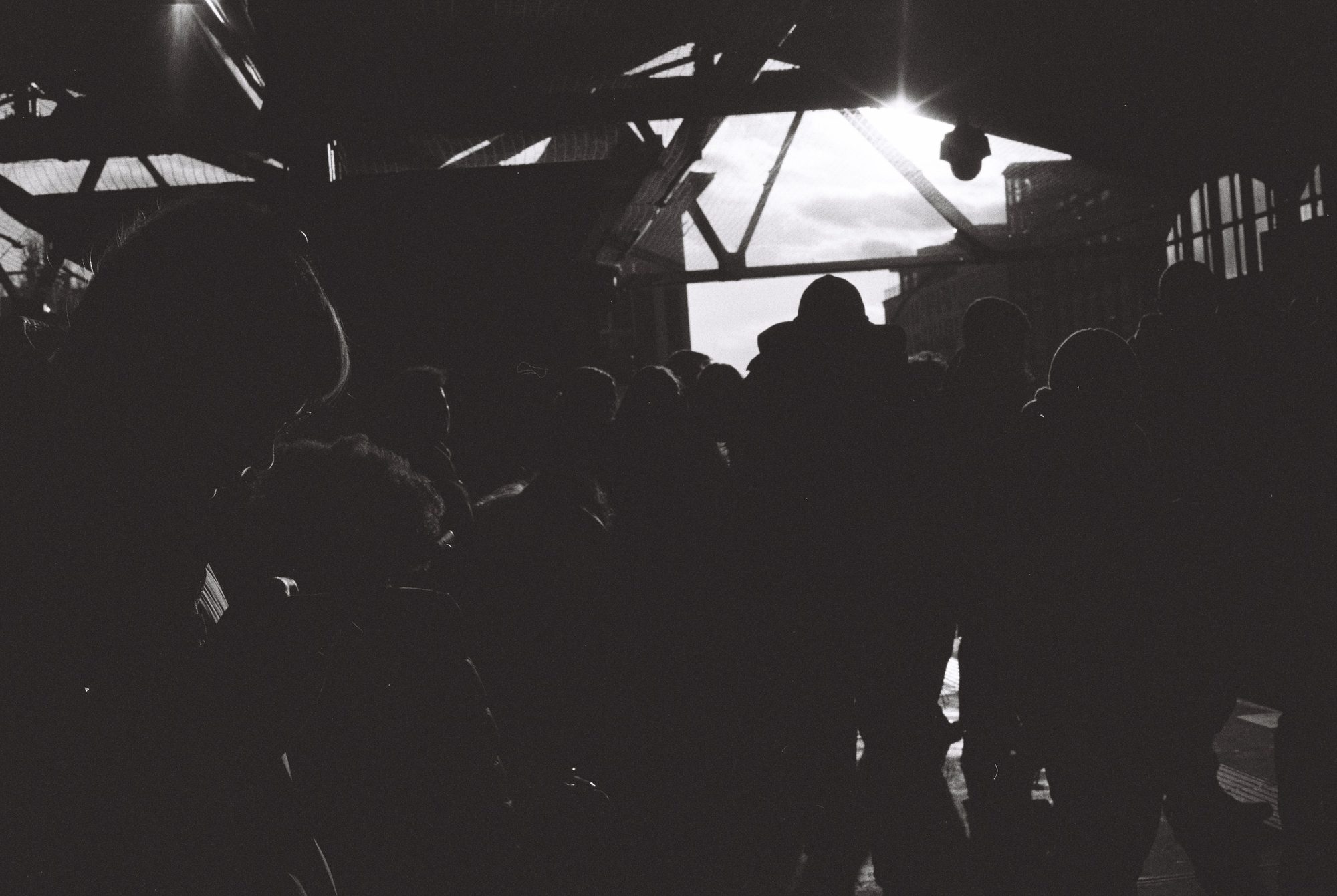 A crowd in silhouette under a steel-framed hall, sun bursting through the roof