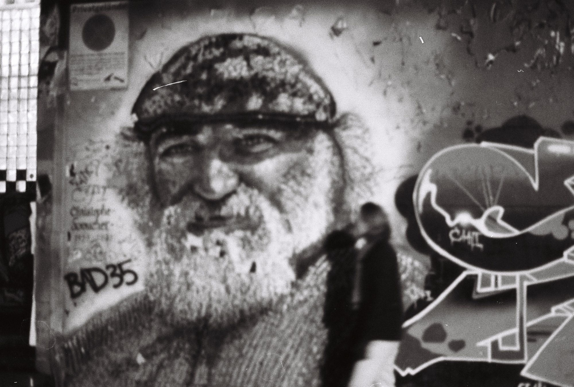 A large mural of a bearded old man in a cap, a blurred passer-by in front