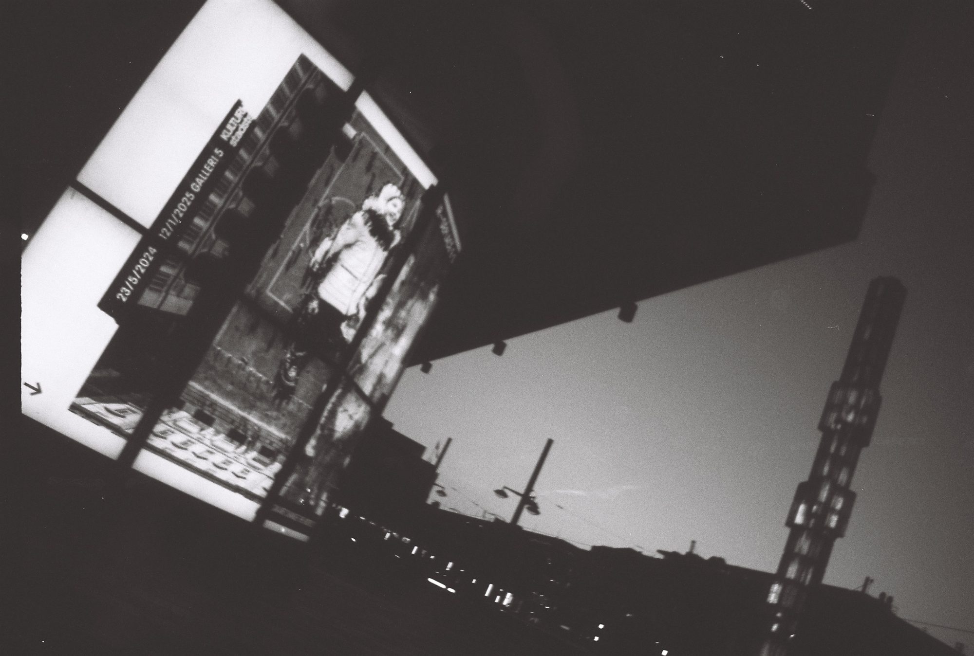 Tilted view of a lit exhibition banner under a dark overhang, skyline and a tower behind