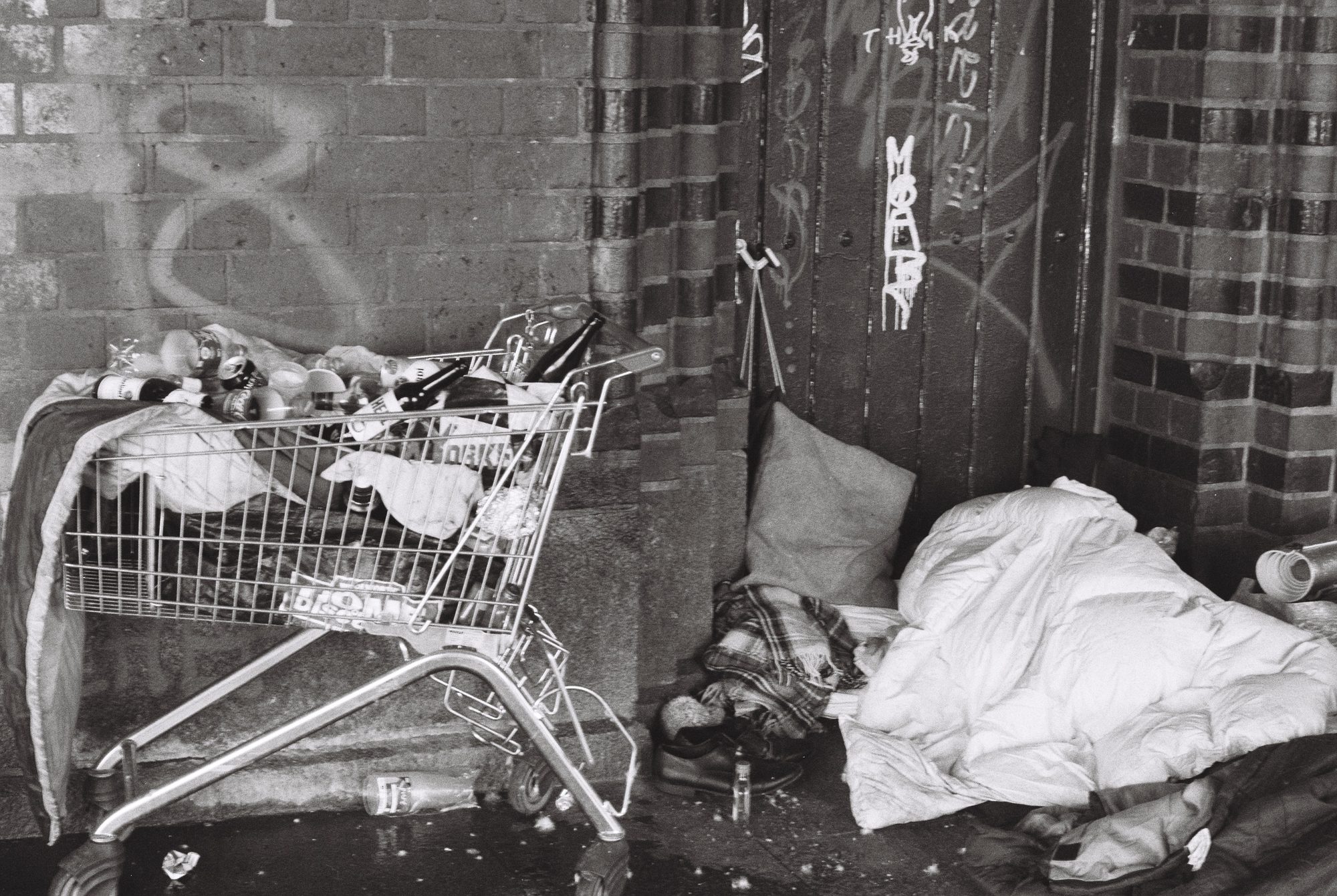 A shopping cart full of bottles beside bedding in a brick doorway with graffiti