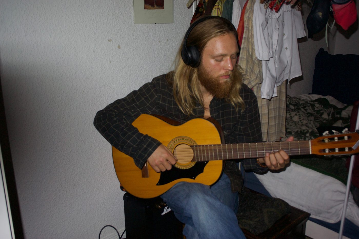 Carl-Fredrik with long hair and a beard, wearing headphones and playing an acoustic guitar
