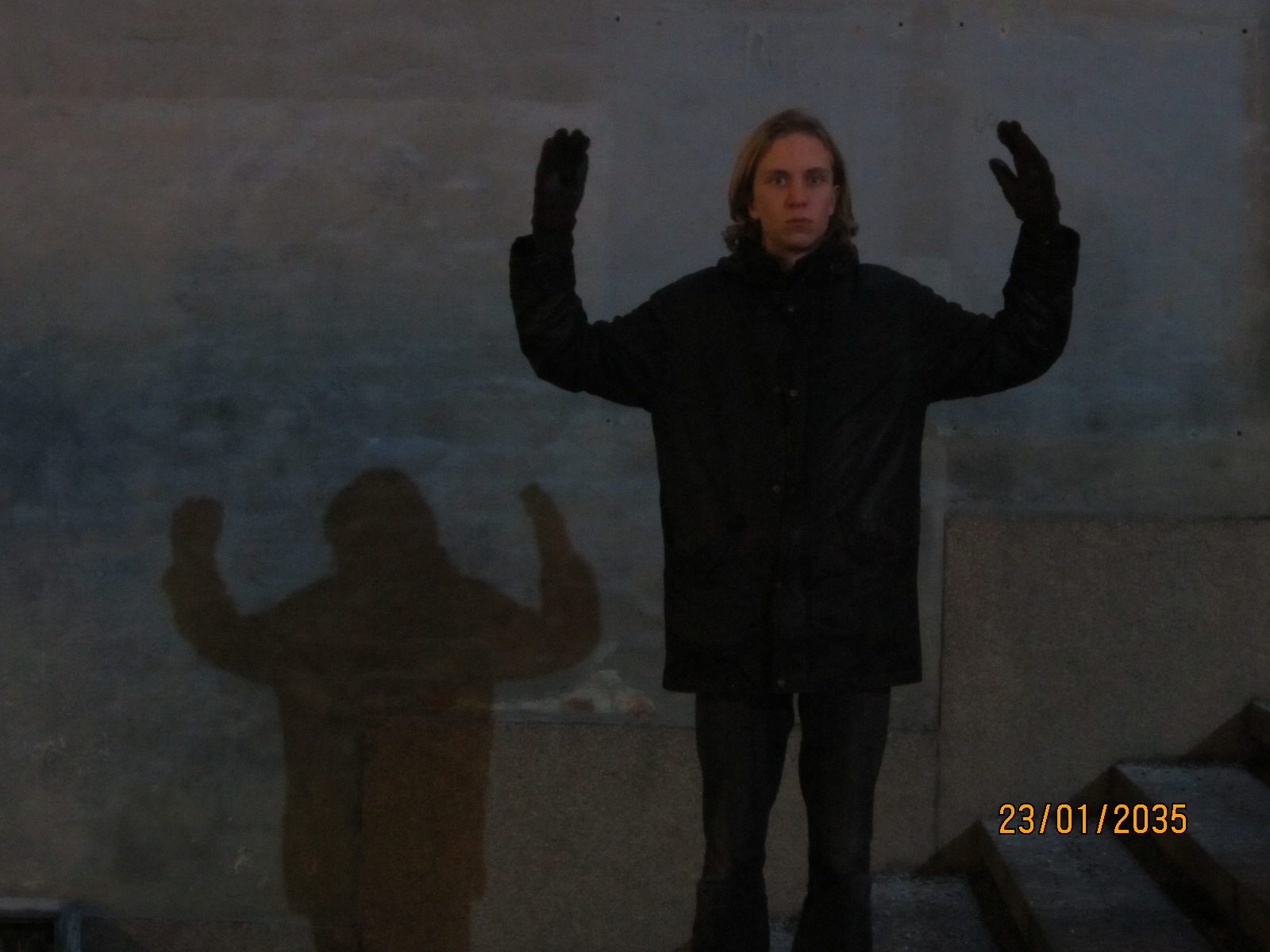 A man in a long black coat with his hands raised against a concrete wall