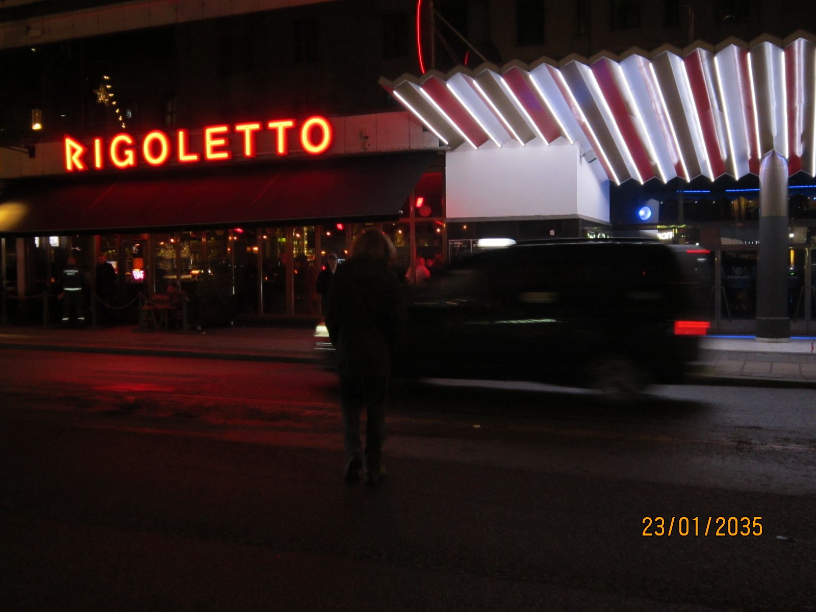 A figure crossing a night street in front of a red neon sign as a car blurs past