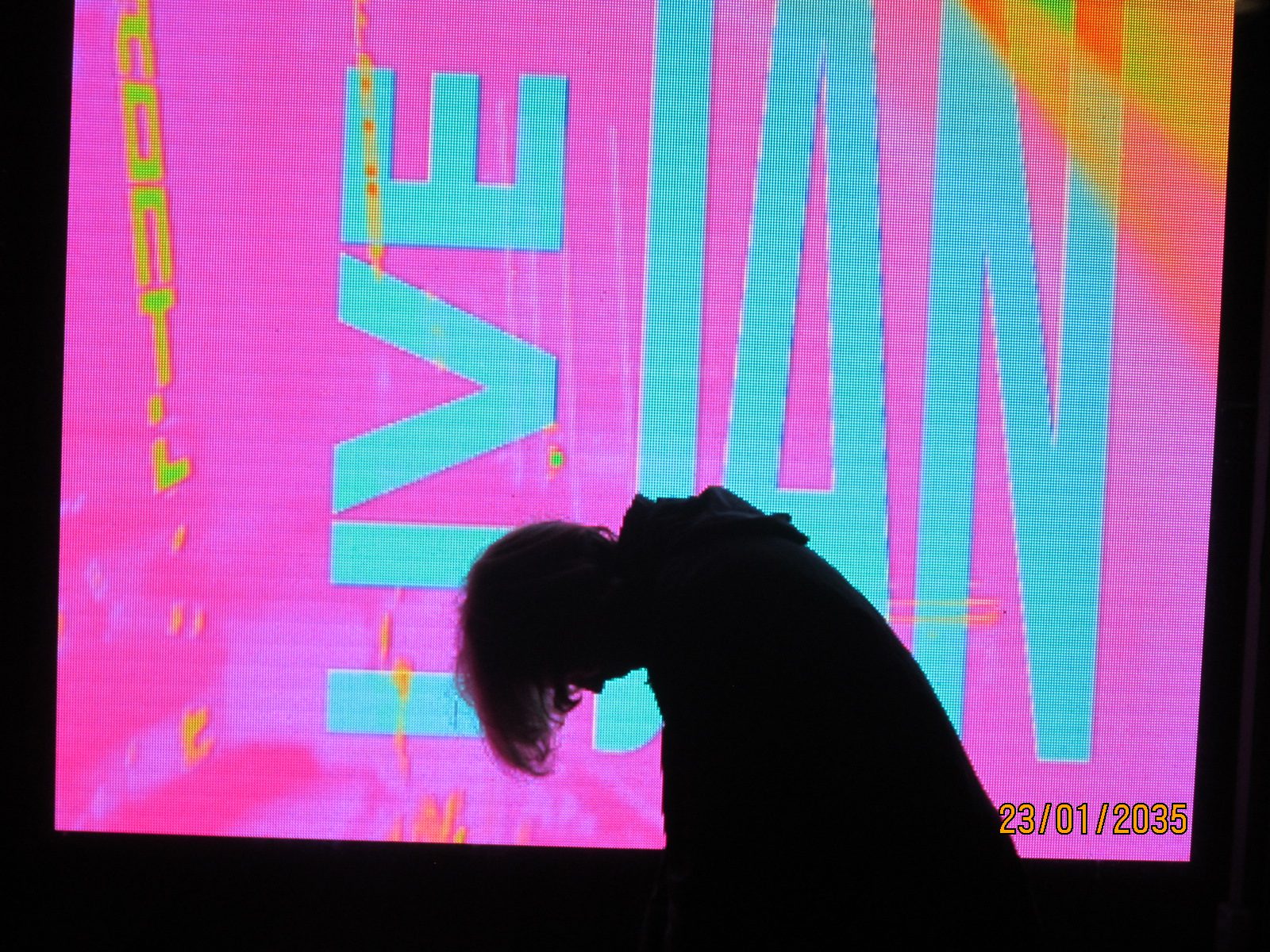 A silhouette bent forward in front of a giant pink and turquoise screen