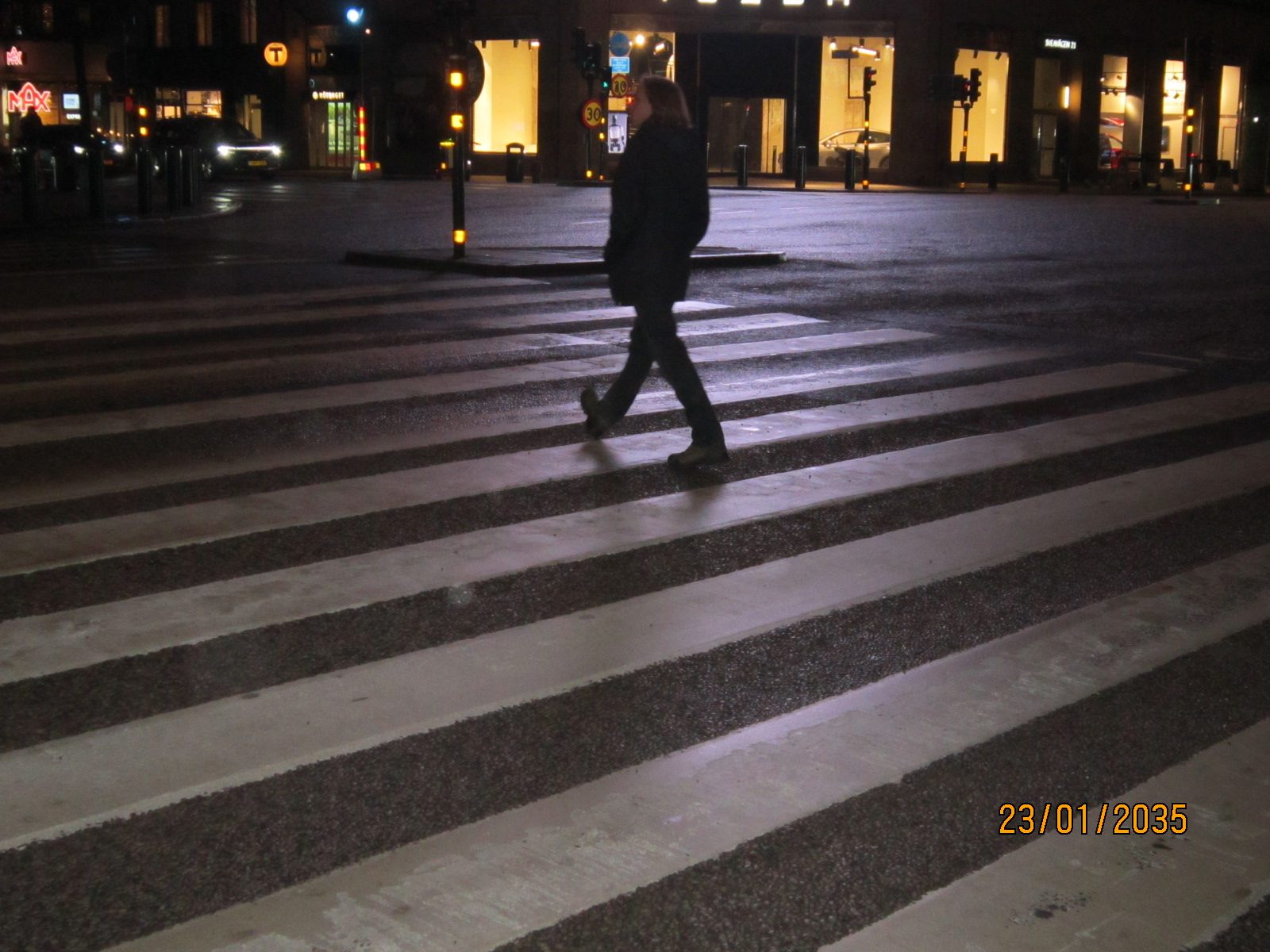 A man in a dark coat walking across a wide zebra crossing at night