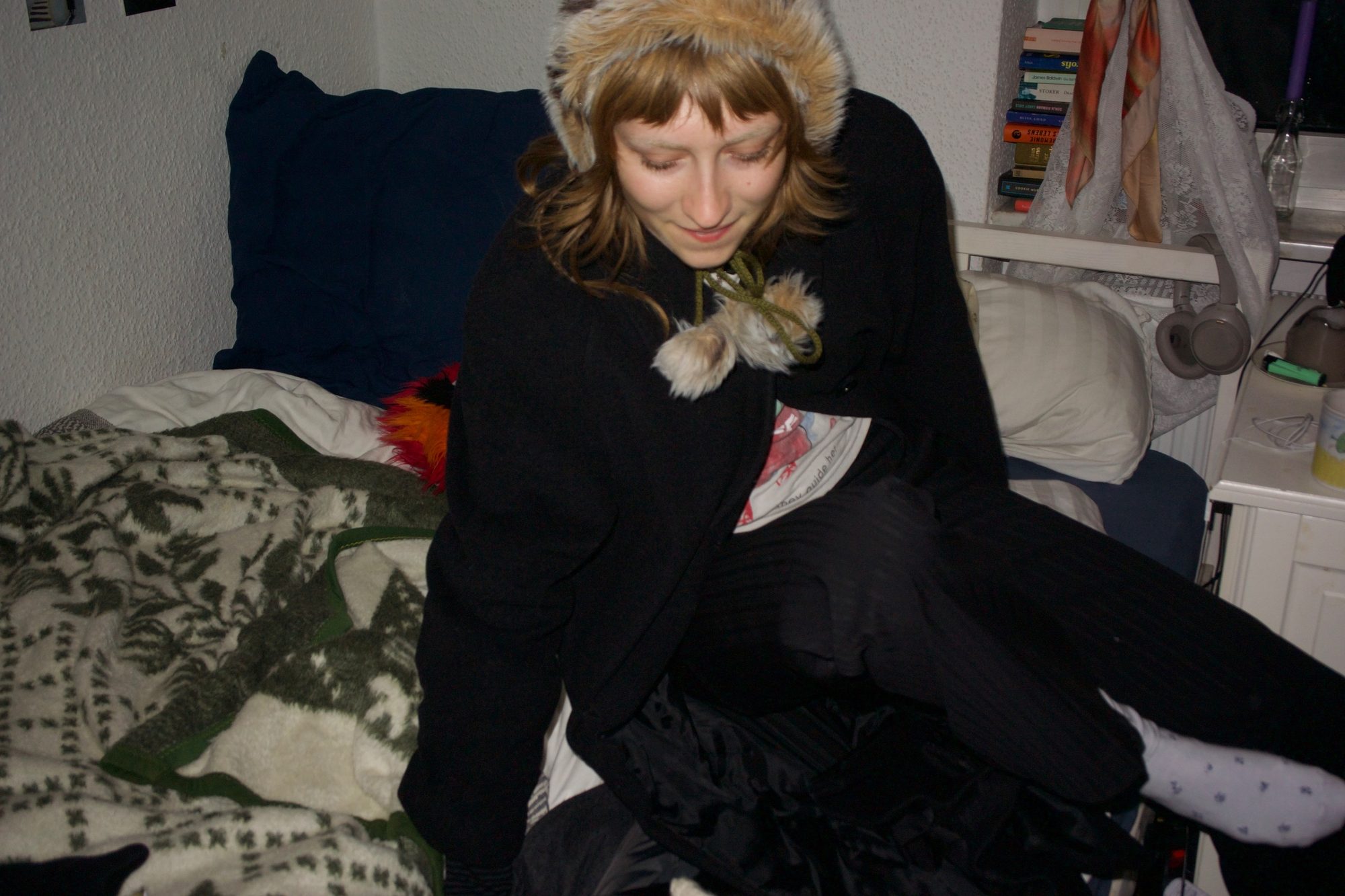 She sits on the bed in a black coat and fur hat, looking down